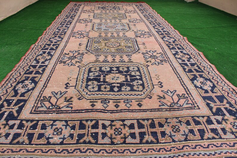 May include: A vintage rug with a pink background and intricate blue, brown, and white patterns. The rug has a geometric design with repeating motifs.