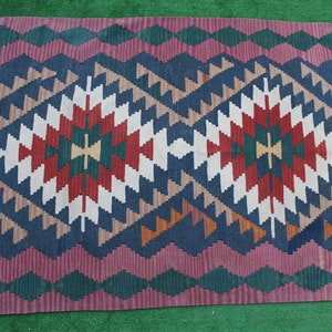 Puede incluir: Una alfombra kilim turca tradicional con un patr&oacute;n geom&eacute;trico en tonos de rojo, azul, verde y blanco. La alfombra tiene un borde con flecos.
