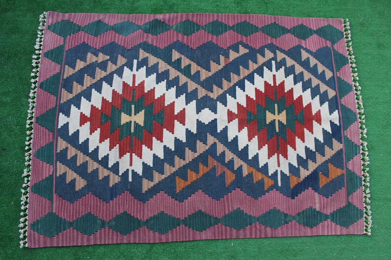 Puede incluir: Una alfombra kilim turca tradicional con un patr&oacute;n geom&eacute;trico en tonos de rojo, azul, verde y blanco. La alfombra tiene un borde con flecos.