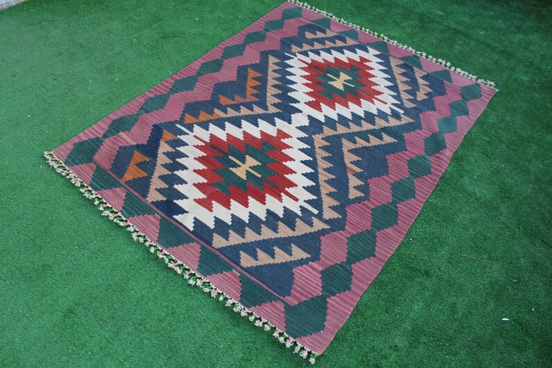 Puede incluir: Una alfombra colorida con un patr&oacute;n geom&eacute;trico en tonos de rojo, verde, azul y blanco. La alfombra tiene un borde de flecos y est&aacute; colocada sobre una superficie verde.