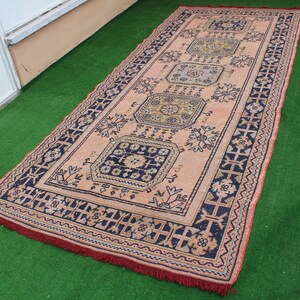 May include: A vintage rug with a pink background and intricate blue and black floral patterns. The rug has a fringed edge.