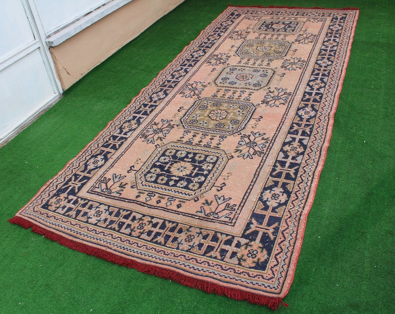 May include: A vintage rug with a pink background and intricate blue and black floral patterns. The rug has a fringed edge.