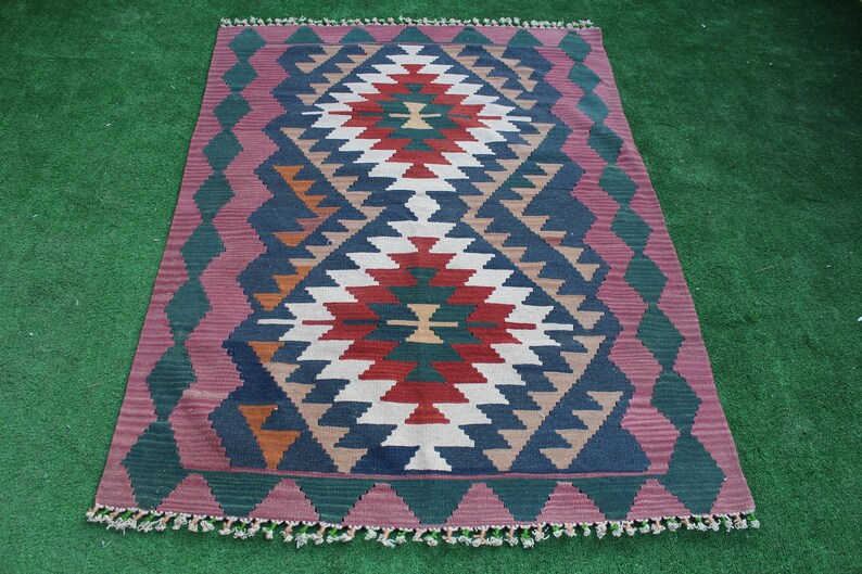 Puede incluir: Una alfombra colorida con un patr&oacute;n geom&eacute;trico en tonos de rojo, verde, azul, blanco y marr&oacute;n. La alfombra tiene un borde de flecos y est&aacute; colocada sobre una superficie verde.