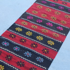 May include: A long, narrow rug with a repeating geometric pattern in shades of red, black, white, yellow, and blue. The rug has a traditional, hand-woven look.