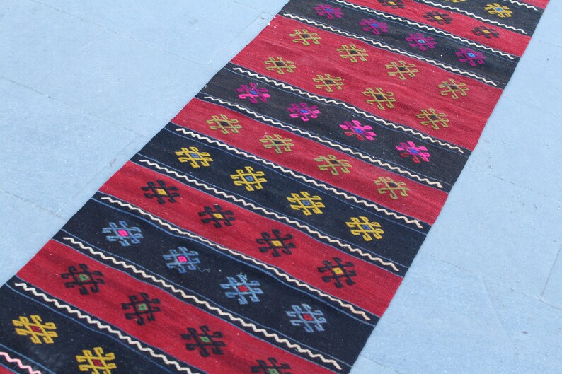 May include: A long, narrow rug with a repeating geometric pattern in shades of red, black, white, yellow, and blue. The rug has a traditional, hand-woven look.