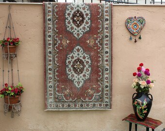 May include: A red, white, and blue patterned rug hangs on a wall with a decorative heart hanging above it. Two hanging planters with red flowers are on either side of the rug.