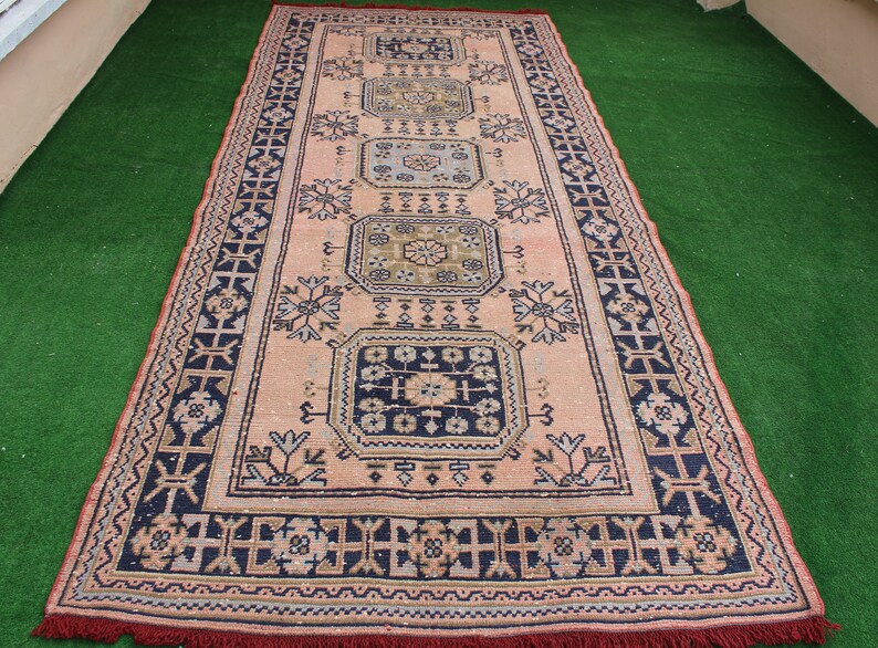 May include: A vintage rug with a pink background and intricate blue, black, and gold patterns. The rug has a fringe border.