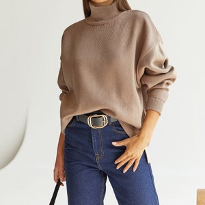 Puede incluir: Un su&eacute;ter de cuello alto marr&oacute;n con mangas largas y un corte holgado. El su&eacute;ter se combina con jeans azul oscuro y un cintur&oacute;n negro con hebilla dorada.