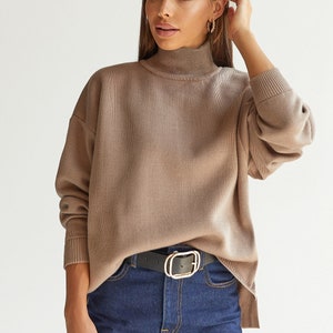 Puede incluir: Un su&eacute;ter de cuello alto marr&oacute;n de corte holgado y mangas largas. El su&eacute;ter se combina con jeans azules y un cintur&oacute;n negro con hebilla dorada.