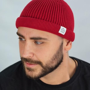 May include: A vibrant red beanie hat with a ribbed knit texture. The hat features a folded brim and a small white tag with black text. The hat is worn by a person with a beard and dark hair.