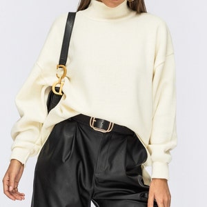 Puede incluir: Un su&eacute;ter blanco de cuello alto con mangas largas y un corte holgado. El su&eacute;ter se combina con pantalones de cuero negro y un cintur&oacute;n dorado.