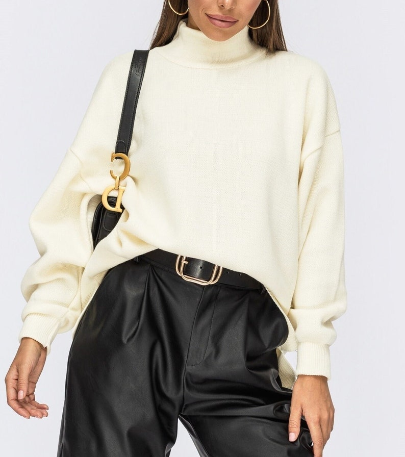 Puede incluir: Un su&eacute;ter blanco de cuello alto con mangas largas y un corte holgado. El su&eacute;ter se combina con pantalones de cuero negro y un cintur&oacute;n dorado.