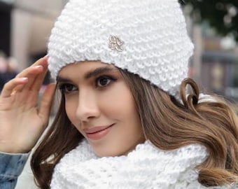 May include: A white knit hat with a faux fur pom-pom and a matching scarf. The hat has a small decorative emblem. The scarf has a faux fur trim. The winter accessories are designed for cold weather.