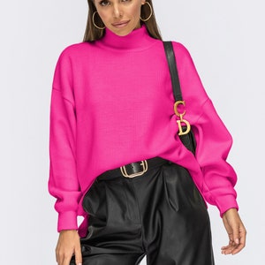 Puede incluir: Un su&eacute;ter de cuello alto de color rosa brillante con mangas largas y un corte holgado. El su&eacute;ter se combina con pantalones de cuero negro y un cintur&oacute;n dorado.