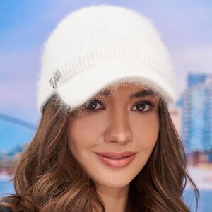 May include: A white fuzzy knit hat with a brim and a small silver embellishment. The hat is worn by a person with long brown hair.