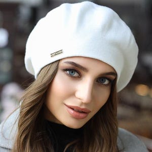 May include: A white beret with a small gold-colored rectangular tag. The beret is made of a soft, textured material. The beret is worn by a person with long brown hair and blue eyes.