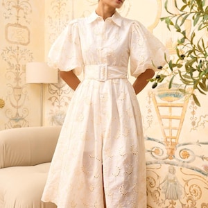 May include: A white eyelet midi dress with a collared neckline, puffed sleeves, and a belt. The dress has a delicate floral pattern and a front slit.