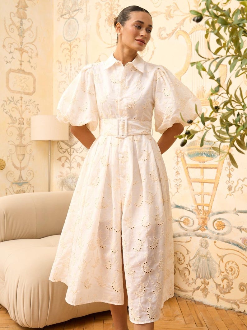 May include: A white eyelet midi dress with a collared neckline, puffed sleeves, and a belt. The dress has a delicate floral pattern and a front slit.