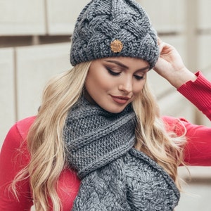 May include: Grey knitted hat and scarf set with a braided pattern. The hat has a small brown leather tag with the word "Handmade" on it.