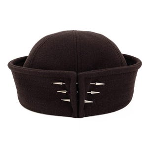 May include: A dark brown felted wool docker cap featuring a rolled brim and a vertical opening at the front. The opening is secured by three pairs of silver-toned metal cone-shaped spikes arranged horizontally on each side of the centre gap.
