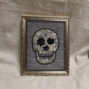 May include: A framed artwork featuring a skull made of white and black buttons on a gray background. The frame is gold with a decorative border.