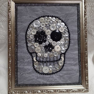 May include: A framed artwork featuring a skull made of white, black, and silver buttons on a gray background. The skull has a single black button for an eye.
