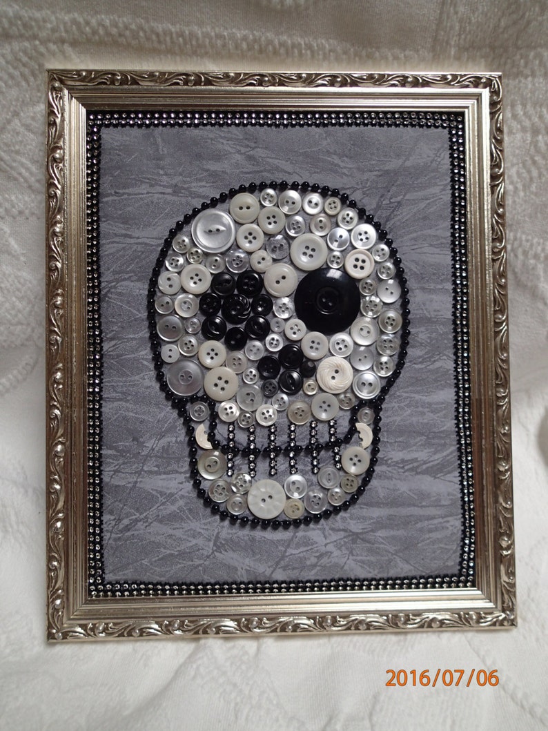 May include: A framed artwork featuring a skull made of white, black, and silver buttons on a gray background. The skull has a single black button for an eye.