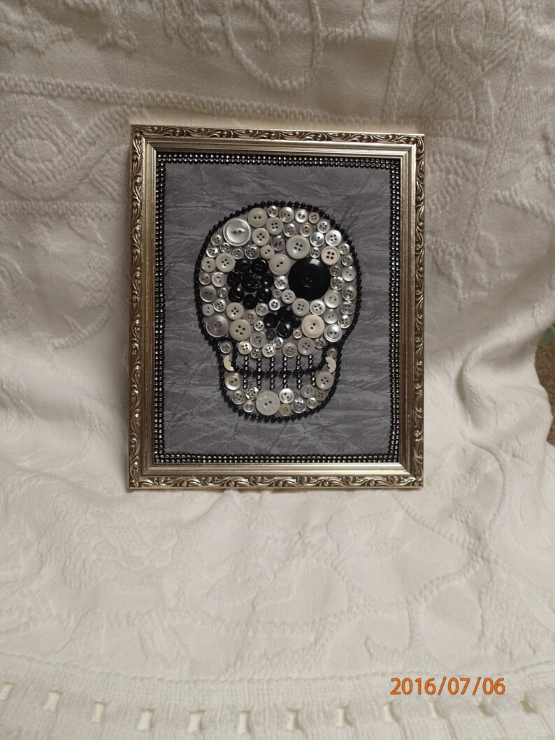 May include: A framed artwork featuring a skull made of white and black buttons on a gray background. The frame is gold with a decorative border.