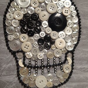 May include: A white skull made of buttons, with black buttons for the eyes and a black button for the nose. The skull is surrounded by a black beaded border.