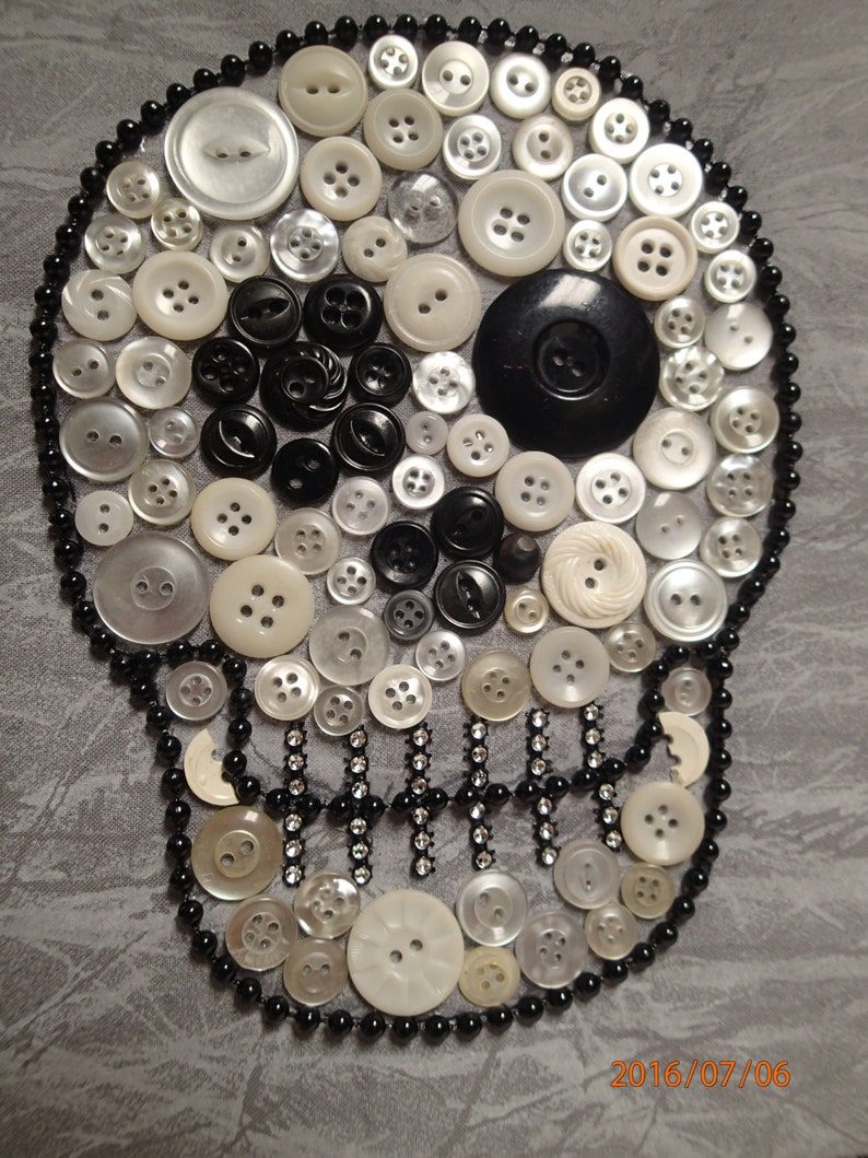 May include: A white skull made of buttons, with black buttons for the eyes and a black button for the nose. The skull is surrounded by a black beaded border.