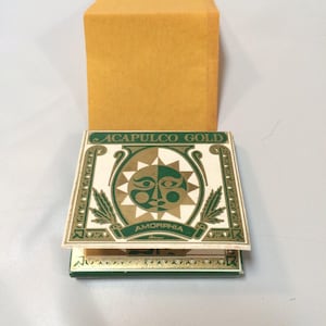 May include: A vintage box with the words "ACAPULCO GOLD" and "AMORPHIA" printed on it. The box has a green and gold design with a sun face graphic. A mustard-colored box is behind it.