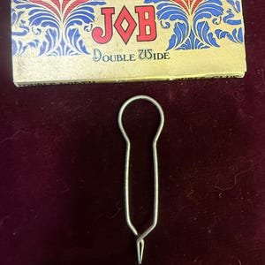 May include: A vintage JOB Double Wide rolling paper pack with a decorative design in red, blue, and gold. A silver metal rolling paper holder is below the pack, with a loop at the top and a pointed end.