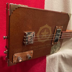 May include: A handmade cigar box guitar with a brown wooden body, metal hardware, and strings. The guitar features the text "STILLWELL STAR ENGLISH No. 2C" and a starburst design. The instrument is set against a red and white background.