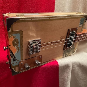 May include: A cigar box guitar featuring a light wood body with the Sobremesa brand logo printed on the front. The instrument includes a chrome bridge, a single black pickup, two silver control knobs, and a wooden neck with metal frets and strings.