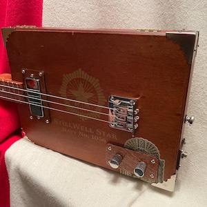 May include: A cigar box guitar with a brown wooden body featuring the text StillWell Star Navy No. 1056. The instrument includes a chrome bridge, a single pickup, two silver control knobs on a wooden plate, and metal corner protectors.