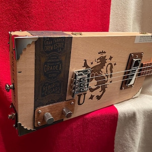 May include: A cigar box guitar featuring a light wood body with a dark blue label reading La Gran Fabrica Drew Estate. The body displays a dark brown lion graphic, a chrome bridge, two silver knobs on a wood plate, and metal corner protectors.