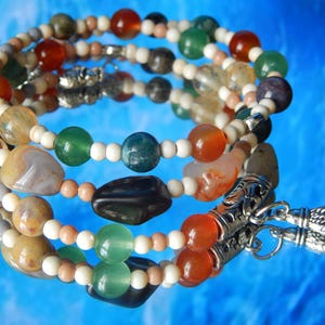 May include: A multi-strand bracelet with a mix of colourful beads and stones. The bracelet includes round beads in orange, green, blue, and white, with larger, irregularly shaped stones. The bracelet is finished with silver-coloured metal accents and a tassel.