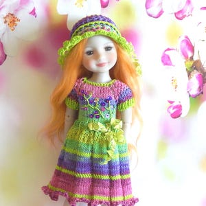May include: A doll with long orange hair wearing a knitted dress and hat. The dress is striped in shades of pink, green, and purple, with floral accents. The hat has a green brim. The doll is posed in front of a soft, blurred floral background.