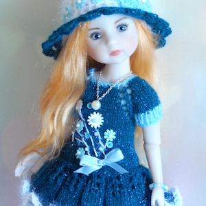 May include: A doll with long, ginger hair wearing a blue knitted dress and matching hat. The dress is decorated with white floral accents and a light blue bow. The hat has a white brim and is decorated with blue flowers.