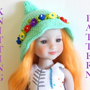May include: A doll wearing a teal knitted hat with a colourful flower trim. The hat has a pointed top and a wide brim. The doll has long, orange hair and is wearing a white dress with a floral pattern.