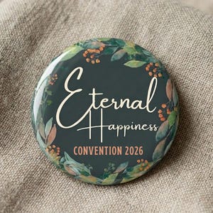 Puede incluir: Un botón redondo con la inscripción "Eternal Happiness" en escritura blanca, rodeado por una corona floral. El texto "CONVENTION 2026" está debajo. El botón tiene un fondo verde oscuro.