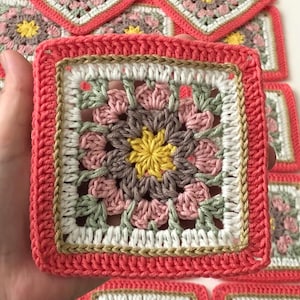 May include: A collection of handmade crochet squares with a floral pattern. Each square has a central yellow flower, surrounded by pink, grey, and green petals, with a white and gold border, and a coral edge. Several similar squares are visible.