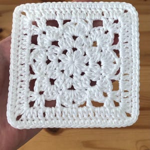 Puede incluir: Un cuadrado de ganchillo blanco. La pieza hecha a mano presenta un diseño floral en el centro y un borde sólido. El cuadrado se sostiene en una mano sobre un fondo de madera. Ideal para proyectos de manualidades.
