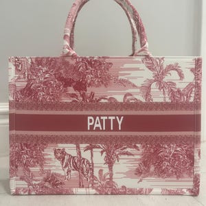 May include: A red and white tote bag with a jungle scene print, featuring a tiger and palm trees. The bag has a red band with the name "PATTY" in white letters. It has a rectangular shape with two handles.