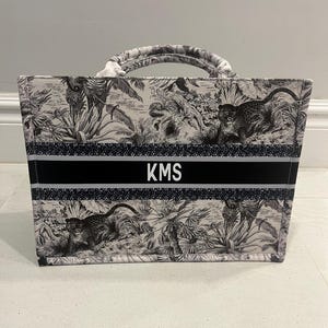 May include: A tote bag with a black and white jungle scene print featuring monkeys and foliage. The bag has a black band with the initials "KMS" in white. The handles are white and black.