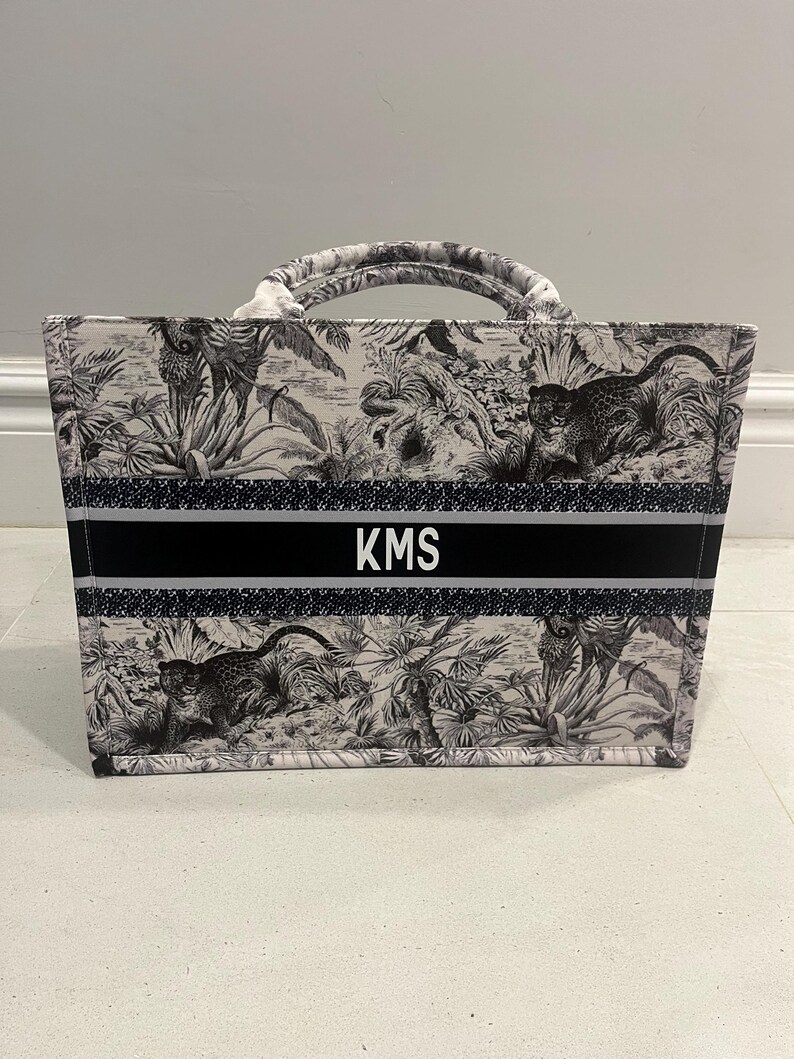 May include: A tote bag with a black and white jungle scene print featuring monkeys and foliage. The bag has a black band with the initials "KMS" in white. The handles are white and black.