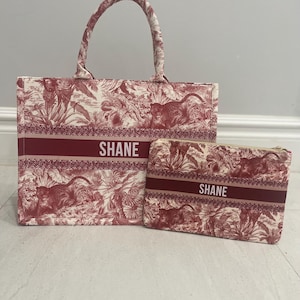 May include: A matching tote bag and pouch set. The bags feature a red and white toile pattern with a jungle theme. The word "SHANE" is printed in white on a red band across both bags. The tote bag has a handle.
