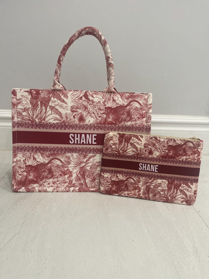 May include: A matching tote bag and pouch set. The bags feature a red and white toile pattern with a jungle theme. The word "SHANE" is printed in white on a red band across both bags. The tote bag has a handle.
