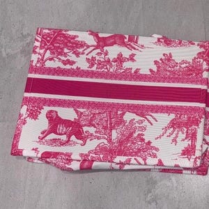 May include: A folded white and pink toile fabric with a repeating pattern of deer, trees, and a tiger. A thick pink stripe runs across the center. The fabric is folded and appears to be a tote bag or similar item.