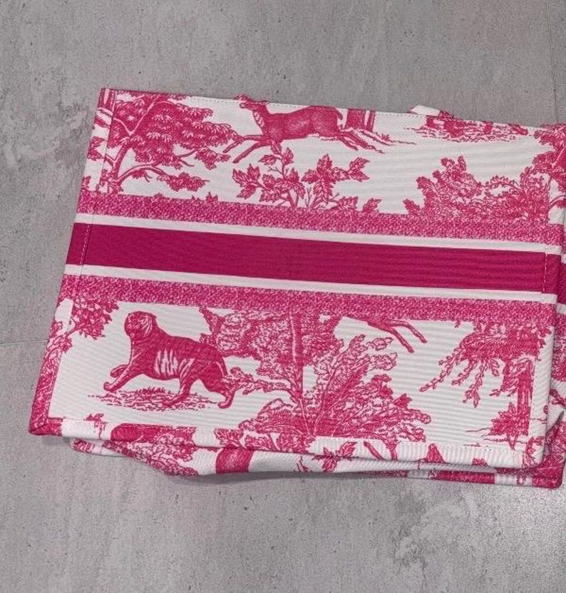 May include: A folded white and pink toile fabric with a repeating pattern of deer, trees, and a tiger. A thick pink stripe runs across the center. The fabric is folded and appears to be a tote bag or similar item.